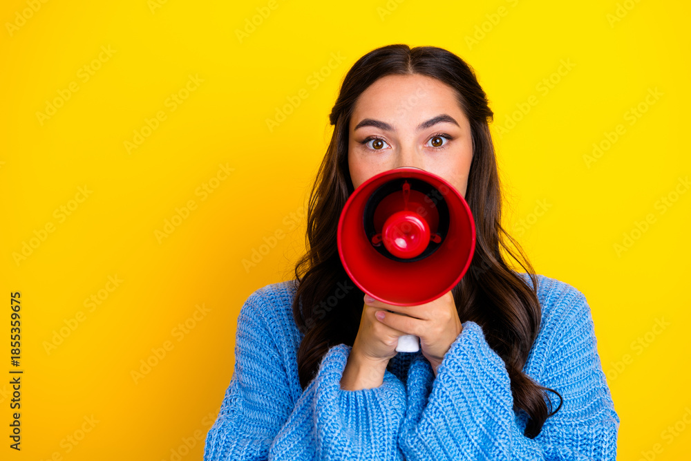 Fototapeta premium Young woman with red megaphone speaks to audience in a bright yellow studio wearing a blue sweater