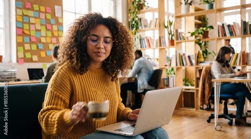 A woman is working in an office with a laptop, holding a cup of coffee. Several other individuals can be seen in the background.