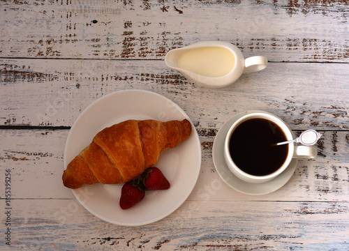 French breakfast, strawberry croissant, and black coffee with cream on a light wooden table.