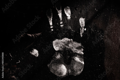 A white palm print contrasts against a pitch black background. The surface appears dusty and scratched, creating a mysterious, eerie, or forensic atmosphere.