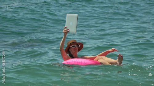 Woman Tablet Ocean. Woman floats on pink ring, raising tablet to capture sunny sea view on summer vacation.
