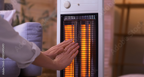 Tableau sur toile Young woman warming hands near electric heater in evening at home, closeup