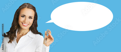 Happy smiling businesswoman or real estate agent showing keys from new house. Portrait of brunette woman at studio, isolated plain blue background with white empty mockup speech bubble