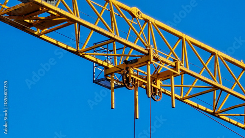 A crane is suspended in the air with a blue sky in the background. The crane is yellow and has a large wheel on the front