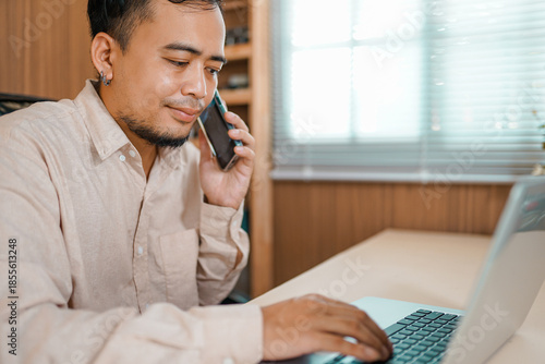 Successful joyful Asian man working inside office at workplace. Businessman talking on mobile phone sitting at desk, working with laptop. 
