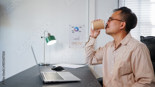 Smiling Asian man drinking coffee at living room, young man having a coffee break in the room.
