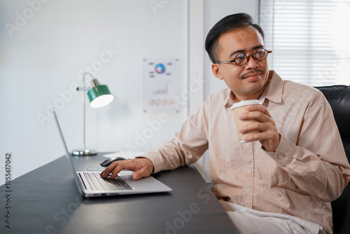 The manager of an Asian man sipping coffee on his desk in a radiant manner.
