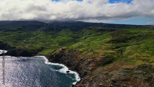 Wallpaper Mural An aerial shot of Maui's rugged coastline with green hills cascading to the ocean, white waves crashing against rocks under a sky with fluffy clouds. Torontodigital.ca