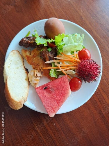 Close up of a white plate containing watermelon chunks, fresh salad, cherry tomato, bread, and tropical rambutan