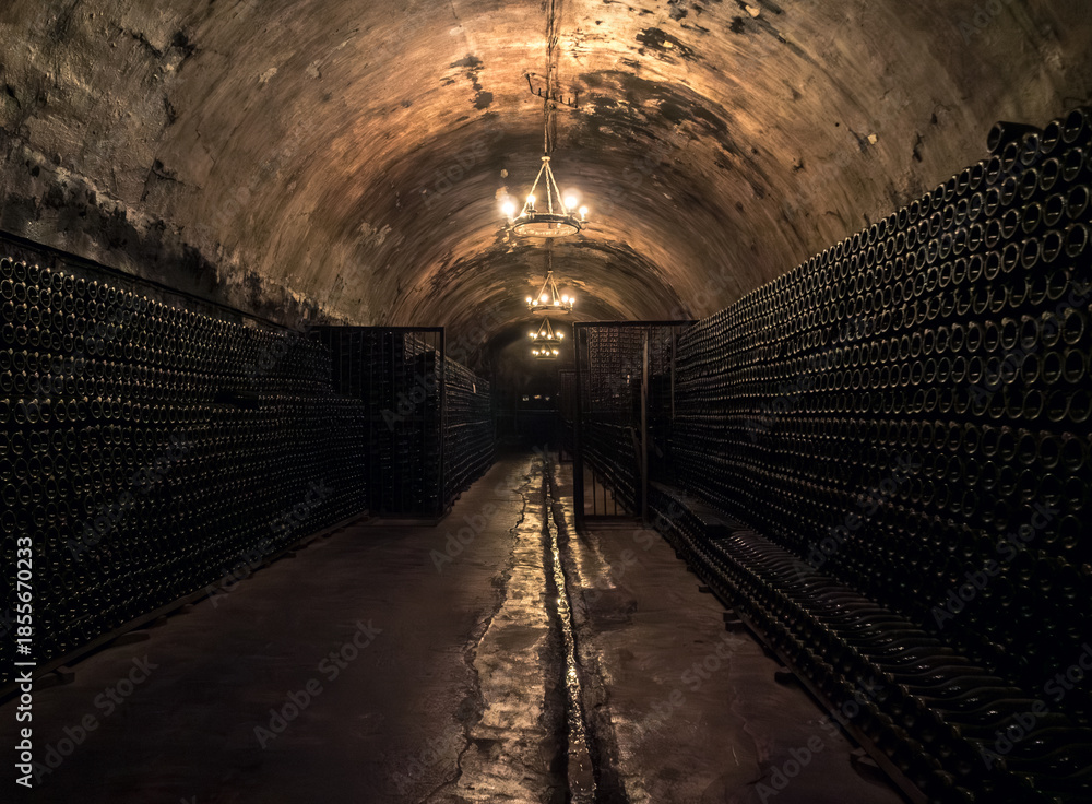 Obraz premium Dark underground wine cellar featuring endless rows of bottles illuminated by soft chandeliers