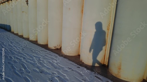 Shadow Of Person On Silo, Snowy Ground And Rusted Pillars, Long Low Sunlight Casting Deep Blue Tones, Silhouette Walking Carrying Shoulder Bag, Slow Contemplative Motion And Cinematic Quiet Mood,