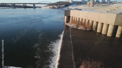 Wide Aerial Dam Spillway And River Channel, Soft Morning Sun On Concrete Pillars Expansive Urban Waterfront View With Repeating Cylindrical Supports, Distant Bridge Arch And City Skyline, White Froth