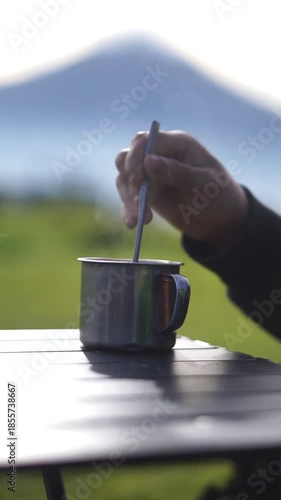 Vertical video footage of a cup of coffee with a folding table on a stretch of green grass outdoors in the open hills, as well as a black folding chair
