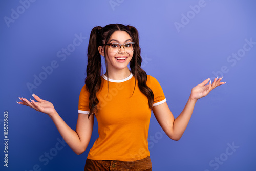Young female student with playful braids wearing an orange shirt stands against a violet blue background smiling and gesturing in a fun lifestyle moment