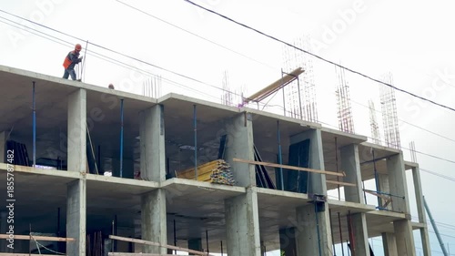 Rooftop construction worker on concrete slab, balancing near edge with crane in background, exposed rebar and concrete columns, scaffolding below, safety helmet visible, dusty