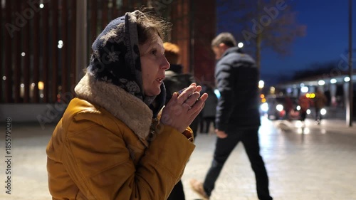 Woman drinks coffee in a public square at night during cold weather near a road with traffic and walking people
