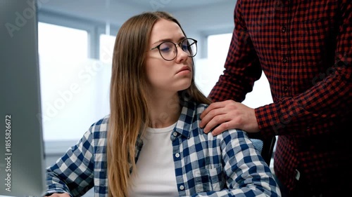 A worried woman works at her computer as a concerned male colleague places his hand on her shoulder in a brightly lit office environment.