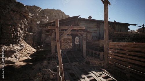 An abandoned western style town showcases rustic wooden buildings and dusty paths under a clear blue sky. The rugged landscape adds to the atmosphere of desolation and history.