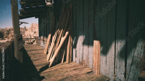 A rustic structure stands in the desert, its wooden porch bathed in sunlight. Stacked planks lean against the weathered wall, reflecting the tranquil yet rugged landscape.