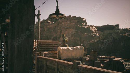 An old western town setting features wooden structures and covered wagons surrounded by rocky terrain. The late evening light casts a warm glow on the rustic buildings and landscape.