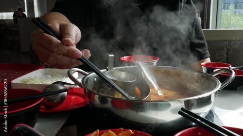 Person eating hot pot shabu shabu with chopsticks and steaming broth in divided metal pot at restaurant table