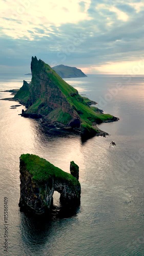 Dramatic rock formations pierce the calm sea off the coast of the Faroe Islands. The warm glow of sunset casts beautiful hues over the landscape, creating a stunning visual experience.