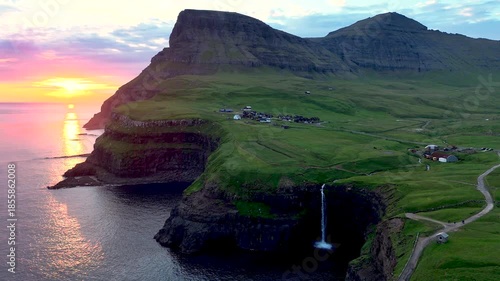 Experience the breathtaking view of Mulafossur waterfall cascading down to the ocean, with the charming Gasadalur village nestled in the lush landscape during golden hour.