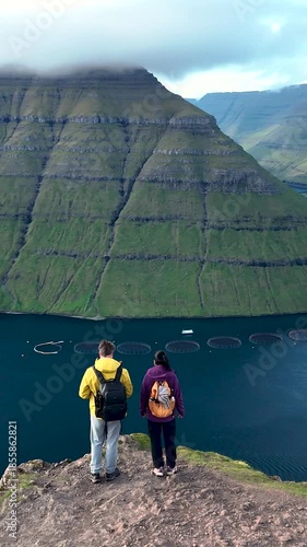 Two adventurers stand on a cliff overlooking stunning blue waters and lush green hills in the Faroe Islands. The serene environment captures the beauty of nature of Klakkur