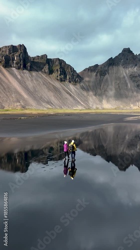 Two visitors trek across the serene black sands of Stokksnes Vestrahorn, surrounded by majestic mountains and mirror-like water reflecting the breathtaking landscape.