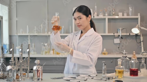 A South Asian woman in a white coat and gloves swirls a flask filled with golden liquid. She works at a perfumery station surrounded by scent strips and aroma bottles