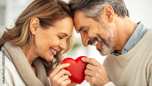 A close-up, tender portrait of a smiling middle-aged couple touching foreheads while jointly holding a small, bright red heart object, symbolizing their deep love and connection.