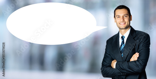 Portrait image - smile young business man wear black dark suit jacket, blue shirt, tie. Businessman standing against blurred office background with white empty mockup speech bubble