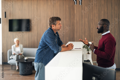 Fotografia Two Men Talk At Reception Desk In Modern Office Lobby, Professional Conversation