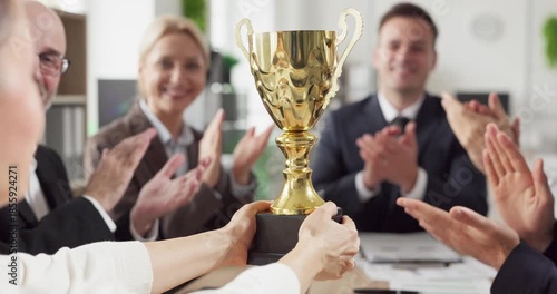 Team business colleagues trophy award success celebration. In a bright office a golden cup is presented as happy coworkers offer applause around the desk. Workplace victory and motivation theme.