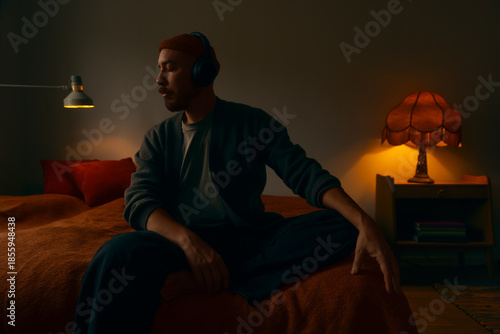 A pensive man wearing headphones and a beanie sitting on a bed in a dimly lit, cozy room. The scene features warm ambient lighting from vintage lamps, creating a moody and cinematic atmosphere