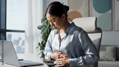 Woman clutching abdomen desk with laptop, holding abdomen office chair with pained expression and tense posture, experiencing stomach pain and discomfort while sitting and working modern office with