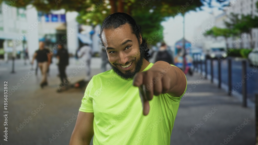 Naklejka premium Man in neon green shirt smiling and pointing finger directly at camera on busy city street; friendly challenge playful.