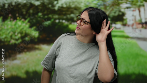 Woman standing in a park wearing glasses and a grey shirt with a hand gesture under the leafy green trees.