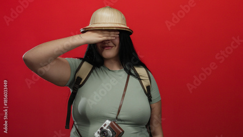 Fotografi Woman saluting with pith helmet and camera around her neck over vibrant red wall