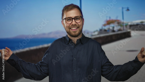 Schilderij op canvas Man smiling with visible face and arms outstretched on a street promenade by the sea, wearing glasses and a dark button shirt; carefree vacation joy
