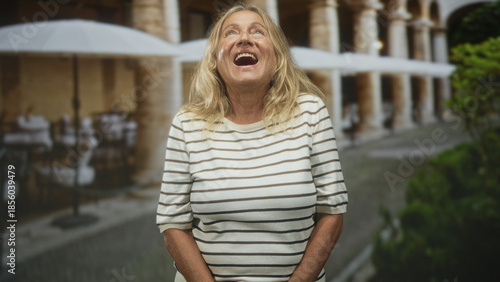 Foto Senior woman with hands clasped looking down in front of building courtyard, striped shirt visible and subtle smile; quiet contemplation