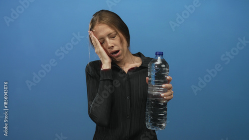 Canvas-taulu Woman holding a large water bottle with hand on cheek yawning in blue studio; tiredness dehydration thirst