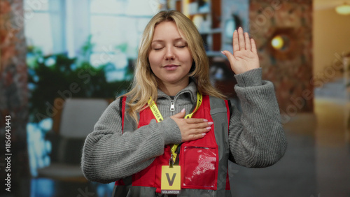 Slika na platnu Woman volunteer in hotel gestures an oath with hand on heart wearing red vest indoors as she expresses commitment and dedication