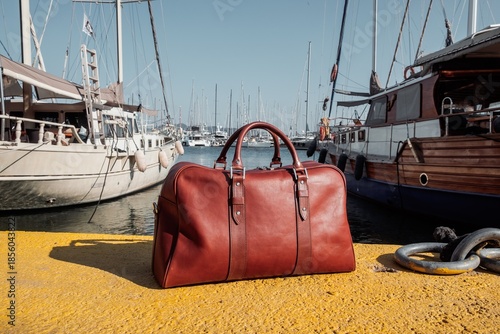 Brown leather duffel bag on a yellow pier with luxury yachts and masts in the background.