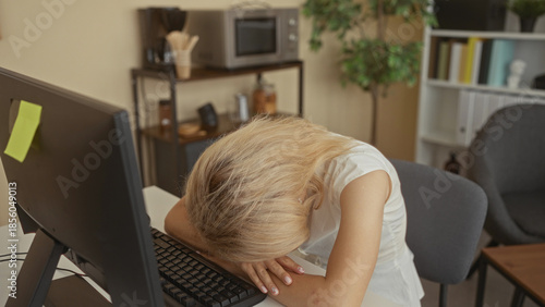 Photos Young blonde woman rests forehead on white desk in modern office building at computer keyboard; tiredness