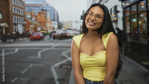 Fotografija Woman smiling and looking up, showing cleavage in a yellow top on a street with storefronts and cars; joy confidence