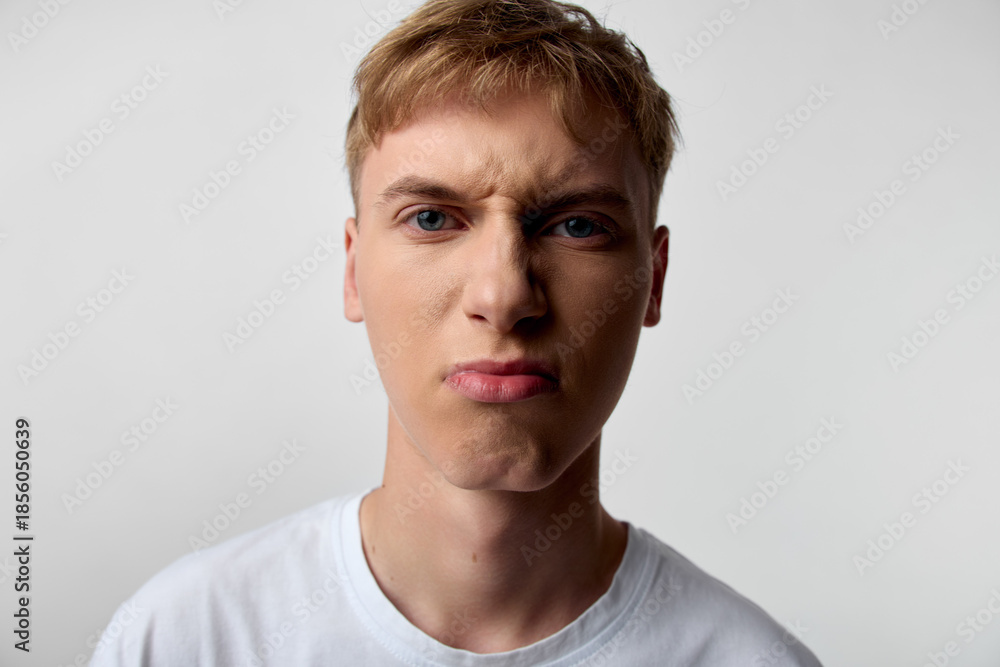 Fototapeta premium Close up of an annoyed man with angry facial expression, pursed lips, and frustrated gaze, wearing a white t shirt against a plain backdrop, conveying irritation, mood, and intense emotion suitable