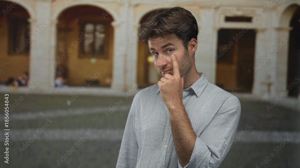 Naklejka premium Man touching eye with finger in building plaza of old town, wearing striped shirt and slight squint; skepticism.