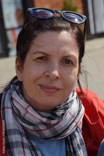 Caucasian brunette woman in red jacket and sunglasses on city street.