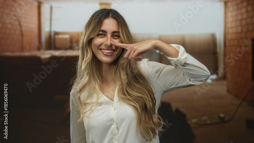 Young blonde woman finger pointing to nose on construction site; playfulness confidence cheerfulness optimism.